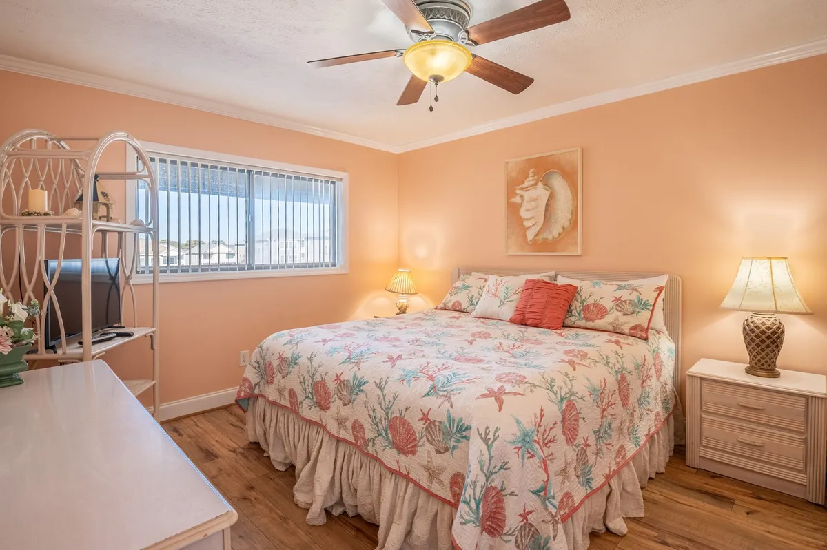 A tranquil bedroom featuring a bed with a coral-patterned quilt, warm wooden floors, a ceiling fan, decorative wall art, and soft lighting from bedside lamps. The space exudes a serene, restful atmosphere ideal for relaxation and unwinding after a busy day.