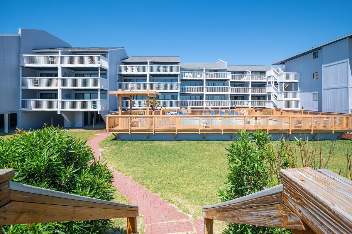 A wide view of the swimming pool area set against the building's facade, with a spacious deck, lounge chairs, and walkways leading to different parts of the property. The scene conveys a cheerful, communal atmosphere.
