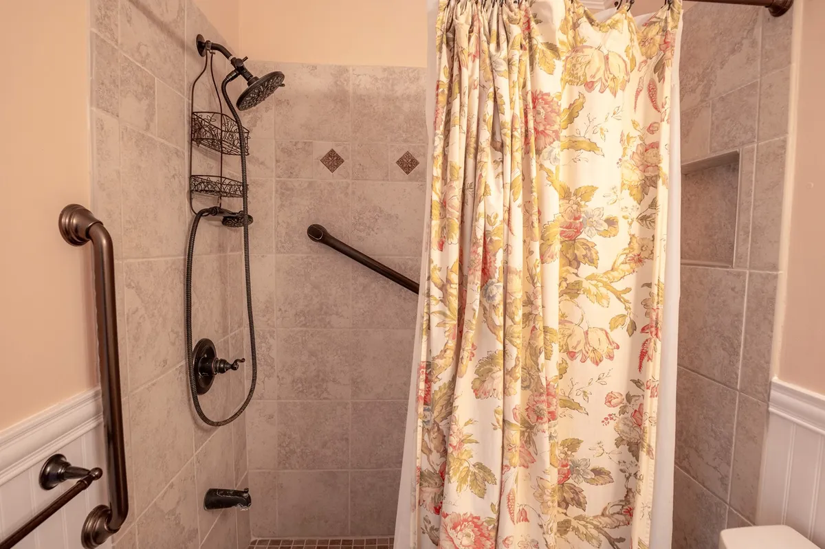A full bathroom with a floral shower curtain, shower head, grab bars, and tiled walls. The space is designed for both style and function, offering a serene spot for self-care routines and rejuvenating showers.