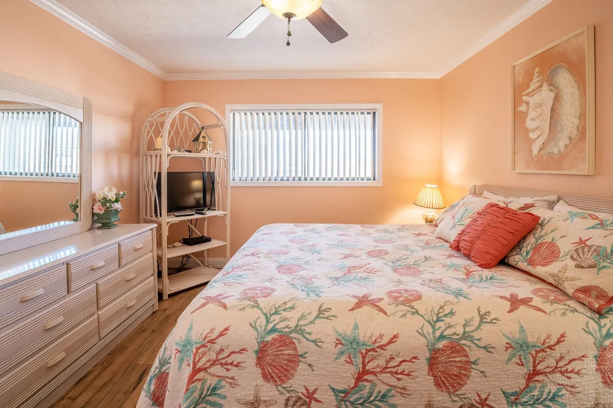 A cozy bedroom with coral-themed bedding, tasteful wall art, a dressing mirror, a television stand, and sunlight streaming through the window. The space feels welcoming and peaceful, inviting guests to relax or enjoy a quiet moment.