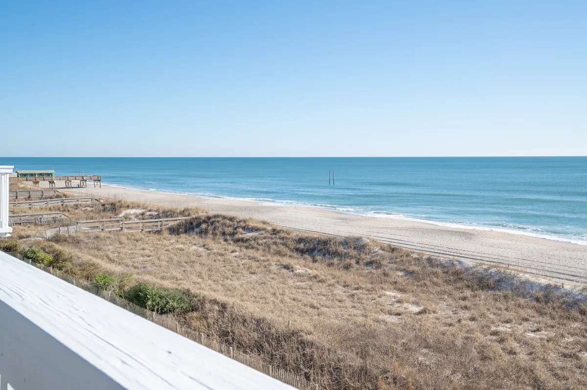 A sweeping view of the beach, ocean, dunes, and a nearby pier sets a classic coastal scene. The wide expanse of sand is perfect for beachcombing, sunbathing, building sandcastles with kids, and watching the waves. The visible dunes and pier anchor the image in a distinct seaside environment, inviting outdoor adventure.