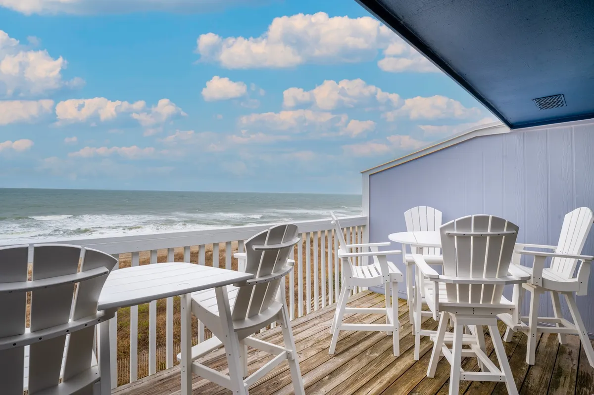 The image showcases a pair of inviting white wooden chairs on a private oceanfront balcony, overlooking the waves of Carolina Beach under a serene, cloudy sky. The tranquil setting is perfect for relaxing, sipping your morning coffee, or sharing sunset cocktails with friends while enjoying panoramic sea views.