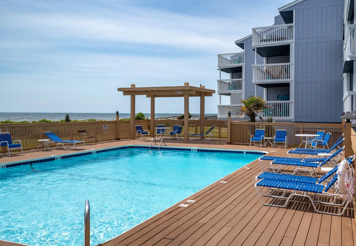 A sparkling swimming pool surrounded by lounge chairs and a pergola, with a clear view of the ocean in the background. Families can be imagined sunbathing, swimming, or gathering for poolside relaxation and social time, all within sight and sound of the waves.