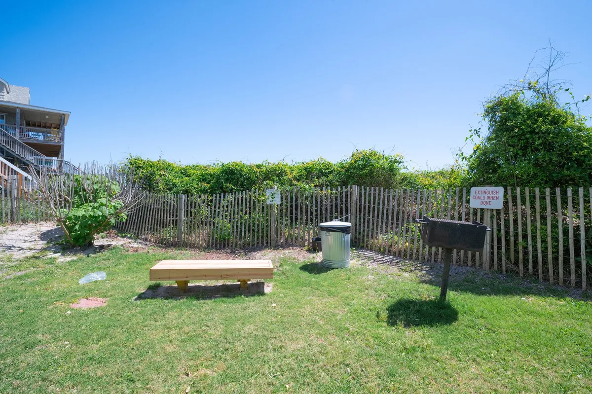 A shaded picnic area nestled among lush greenery, featuring a picnic table, benches, a grill, and a nearby trash can. Sunlight filters through the foliage, creating an inviting space for outdoor meals and gatherings.