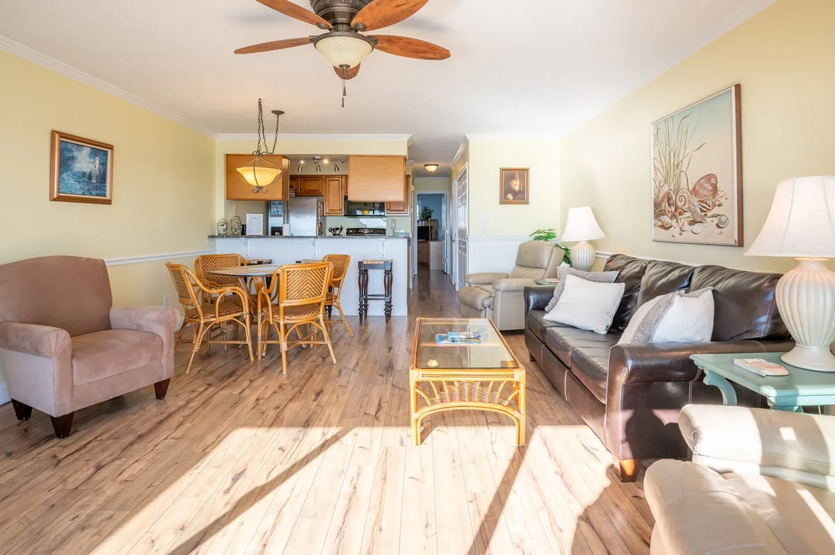 This image features a comfortable sofa, a dining table, a coffee table, and a ceiling fan, all arranged in a spacious shared living area. The room is bathed in natural light and designed for casual gatherings, dining, and relaxation, making it an ideal hub for friends or groups to socialize and unwind together after a day at the beach.