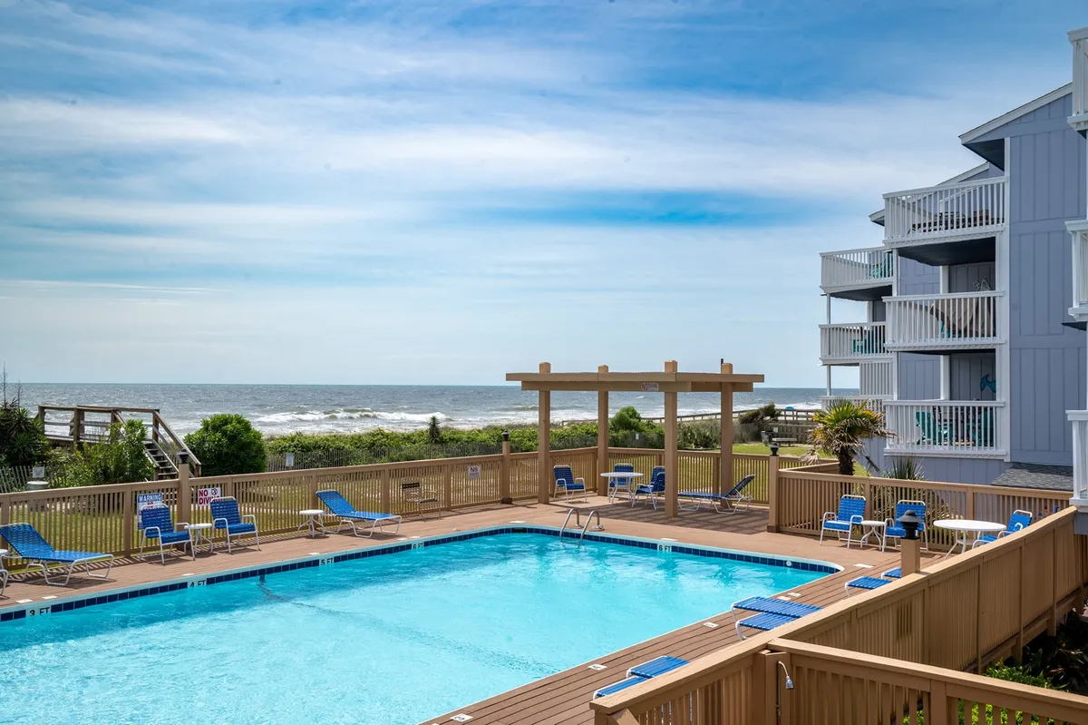 A sun-drenched oceanfront swimming pool surrounded by deck chairs and a wooden pergola, with the shimmering ocean visible in the background. Guests can be seen relaxing poolside, enjoying the inviting atmosphere.